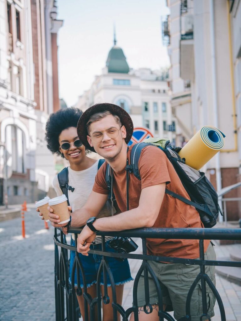 Young couple with backpacks standing on a railing in a city, looking for cheap flights for their next adventure.