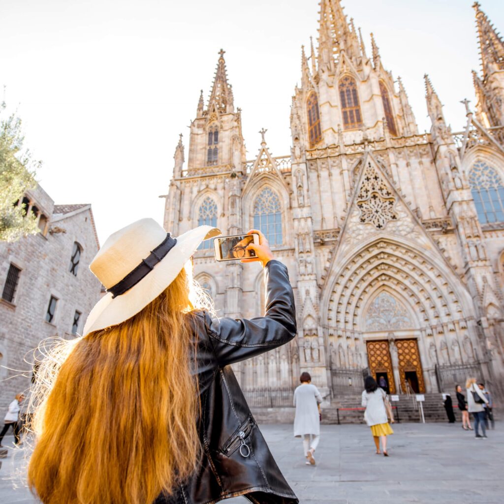 In Barcelona, Spain a woman in a hat takes a picture in front of a cathedral on a walking tour during her holiday.