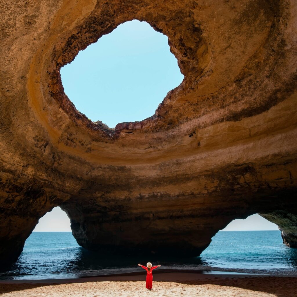 A woman in a red dress standing inside a unique cave with an opening at the top on a beach in Portugal, enjoying a holiday she managed to book with cheap flights through LetsFly.co.uk