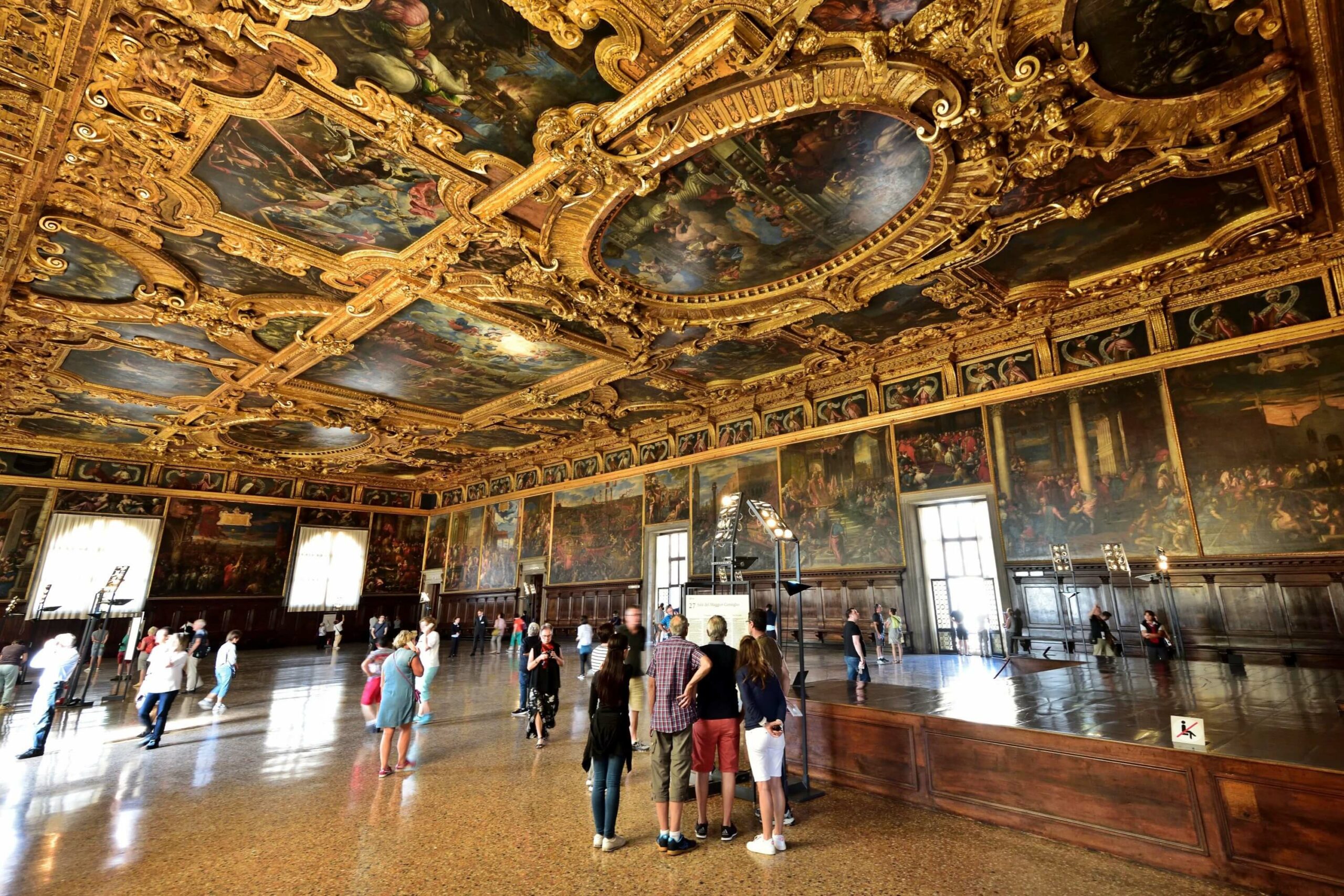 A group of people standing in a large room inside the Venice Doge Palace with ornate ceilings.