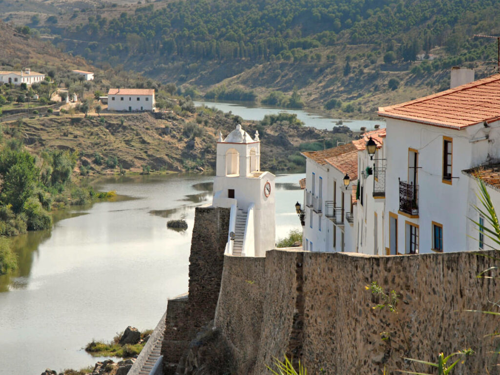 Wall of an old moorish fortress with a village of white houses inside, surrounded by a peaceful river and golden hills