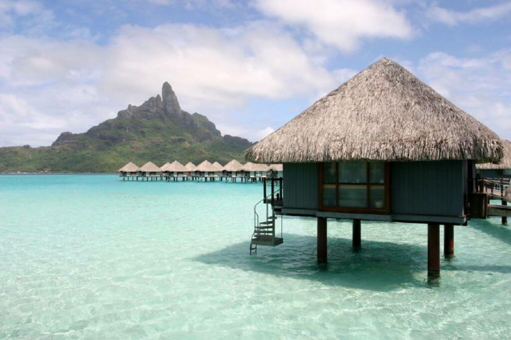 overwater house standing on beautiful beach with blue and crystal clear waters and a green mountain in the background
