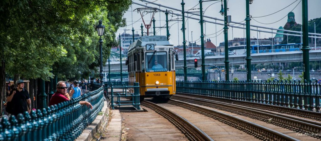 yellow electric tram in Budapest, surrounded by an iron fence, trees and people looking at the streets